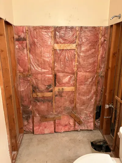Bathroom stripped to studs with pink insulation, plastic vapor barrier and exposed drain in the floor