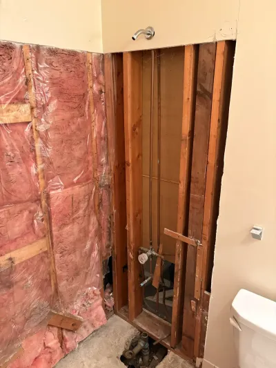 Bathroom shower stripped to studs and insulation, with exposed copper piping, floor drain and chrome shower arm.