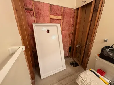 Bathroom stripped to studs and pink insulation with a white shower pan leaning against the framing.