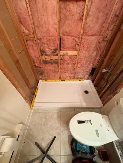 White shower base being leveled inside a bathroom alcove stripped to studs and pink insulation.