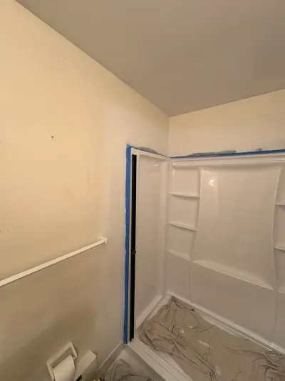 Bathroom mid-remodel with a white fiberglass shower surround masked in blue tape and drop cloths on the floor.
