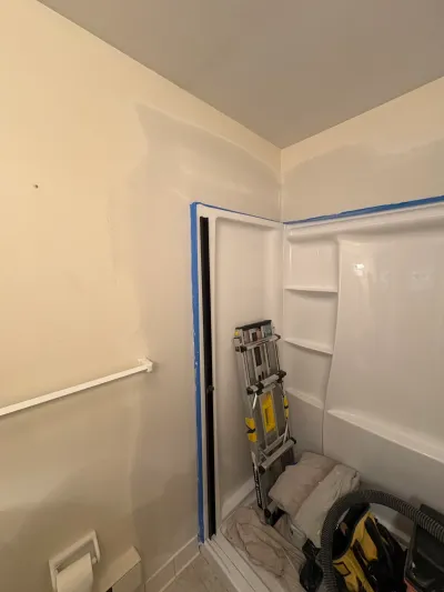 Bathroom mid-renovation with taped-off white tub surround, patched drywall, ladder and shop vacuum inside.