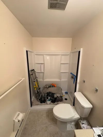 Bathroom under renovation with white shower surround holding a ladder, tools and paint can beside a toilet.
