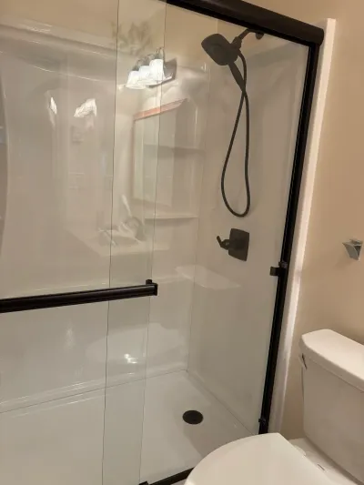White acrylic alcove shower with bronze-framed sliding glass doors and handheld showerhead beside a white toilet.
