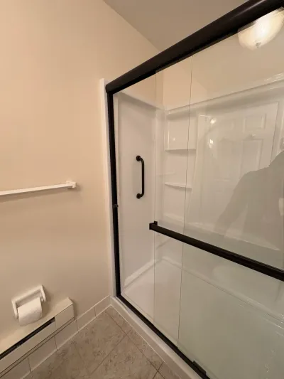 White acrylic alcove shower with bronze-framed sliding glass doors, beige walls and tile floor.