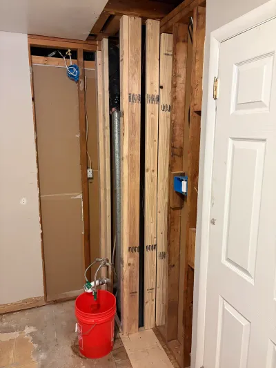 An open closet under renovation showing exposed studs, ductwork, plumbing valves, and a red bucket on the floor.