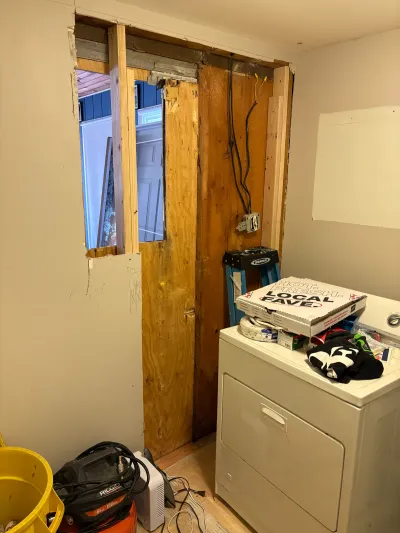A laundry room wall is cut open during remodeling, exposing framing, wiring, and the exterior beyond.