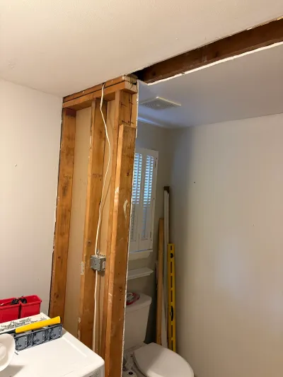 Bathroom under renovation with exposed wall studs, wiring, and an opening leading to a room with a toilet.