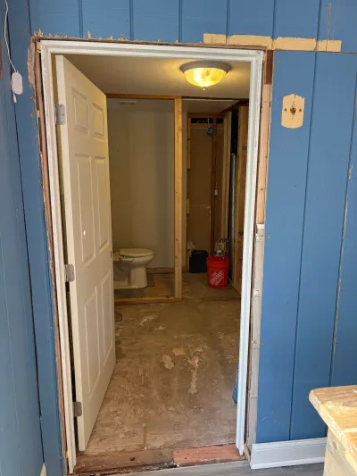An open bathroom doorway reveals exposed framing, a toilet, and a bare subfloor mid-renovation.