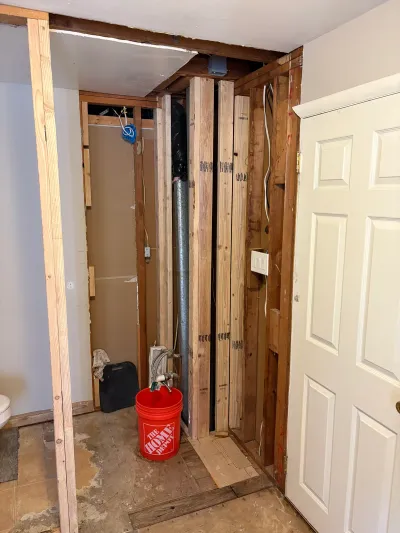 Bathroom under renovation with exposed wall studs, ductwork, and wiring, plus a bucket of tools on the floor.