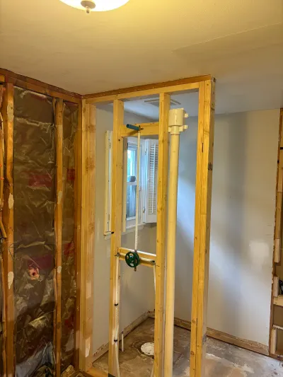 Bathroom under renovation with new wall framing, exposed insulation, and plumbing pipes around a window.