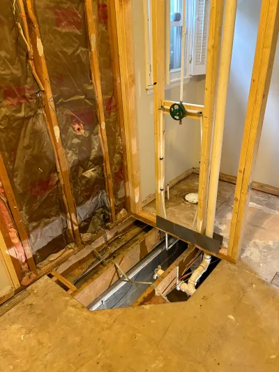 Bathroom under renovation with exposed wall studs, insulation, and a cut-open subfloor revealing plumbing and ductwork.