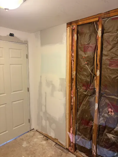 Room mid-renovation with exposed wall studs, insulation, and rough electrical wiring next to a patched drywall wall.