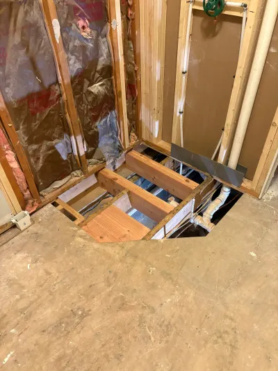 Bathroom subfloor cut open to expose joists, plumbing rough-in, and wall framing during renovation.