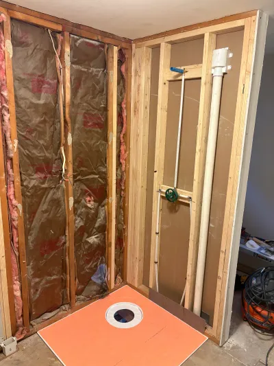Bathroom shower under construction with exposed insulation, framing, and rough-in plumbing.