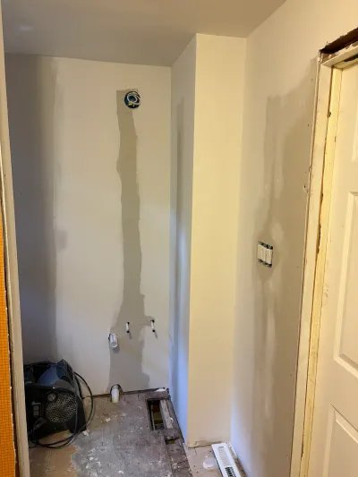 A bathroom mid-renovation with taped drywall seams, exposed subfloor, and a drying fan on the floor.