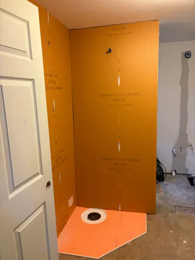 Shower stall mid-renovation with orange waterproofing board installed and drain rough-in visible.