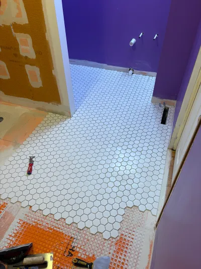 Bathroom floor mid-installation with white hexagon tile over orange waterproofing membrane and rough plumbing visible.