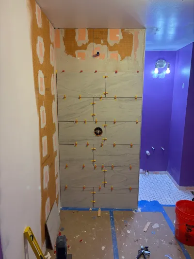 A bathroom shower wall mid-installation with large porcelain tile, exposed waterproofing membrane, and rough plumbing visible