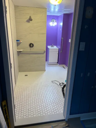 Newly tiled accessible bathroom with a curbless shower, hex tile floor, and purple walls, seen through an unfinished doorway.