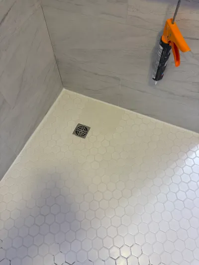 Close-up of a shower corner with white hexagon tile flooring and grey tile walls, caulk gun hanging above.