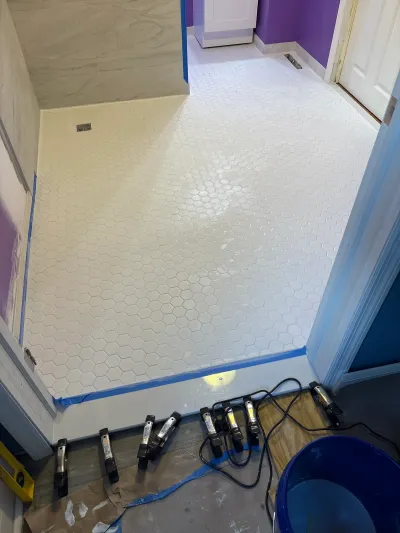 Bathroom under renovation with new white hexagon tile floor and work lights staged near the shower entry.
