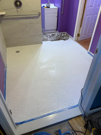 Bathroom mid-renovation with white hexagon tile flooring, marble-look shower walls, and grab bars installed.