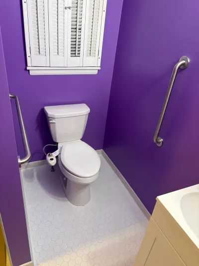 A toilet nook with vivid purple walls, hex tile flooring, and stainless grab bars for accessibility.