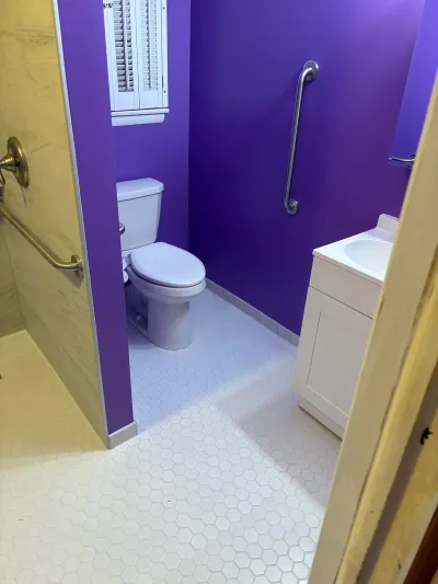 A small bathroom with purple walls, hexagon tile floor, a curbless shower, toilet, and white vanity.