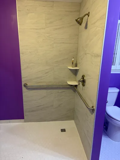 Curbless walk-in shower with marble-look tile, gold fixtures, and grab bars, framed by a purple accent wall.