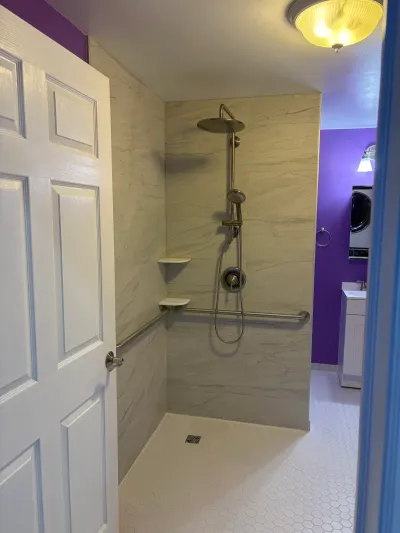 Walk-in shower with rainfall head, grab bars, and tile walls, seen through an open door into a laundry area.