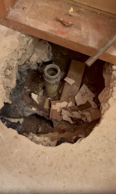 A demolished section of bathroom concrete flooring reveals a drain pipe and construction debris.