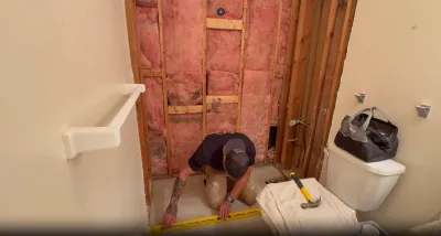 Worker measuring a bathroom subfloor beside an exposed stud wall with insulation during a remodel.