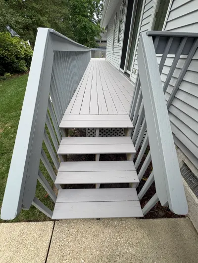 A grey painted wood deck with railings leads down a staircase to a concrete walkway beside the house.