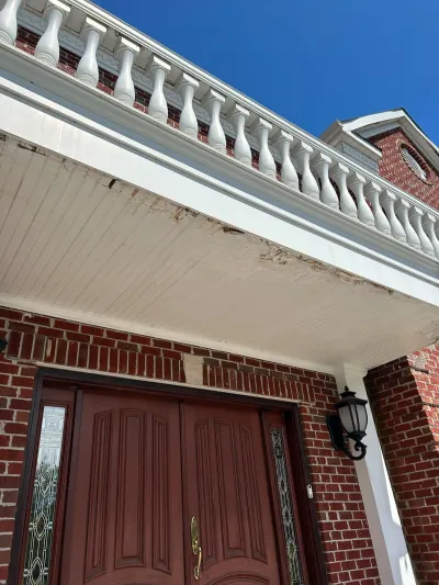Close-up of a damaged white porch ceiling with peeling paint beneath a brick home's balustrade and entry door.