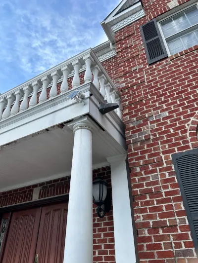Close-up of a brick home's white front portico columns and balcony railing with some visible brick cracking.