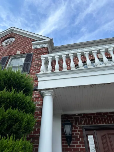 Low-angle view of a brick home's white balcony railing, column, and front porch with visible paint wear.
