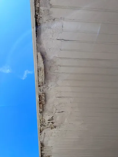Close-up of a badly cracked and crumbling stucco edge along a roofline, seen against a blue sky.