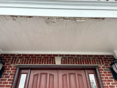 Close-up of a porch soffit with peeling paint above a red brick entryway with double doors.
