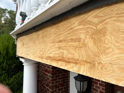 Close-up of new plywood fascia board being installed under a porch roof with white railings and brick below.