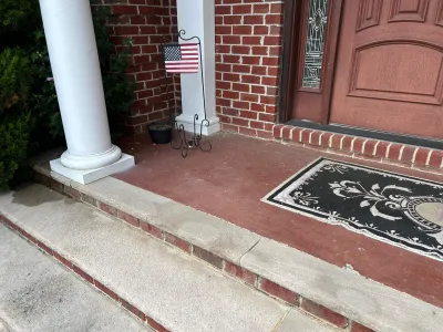 Brick front porch with a white column, decorative door, American flag stand, and a welcome mat.