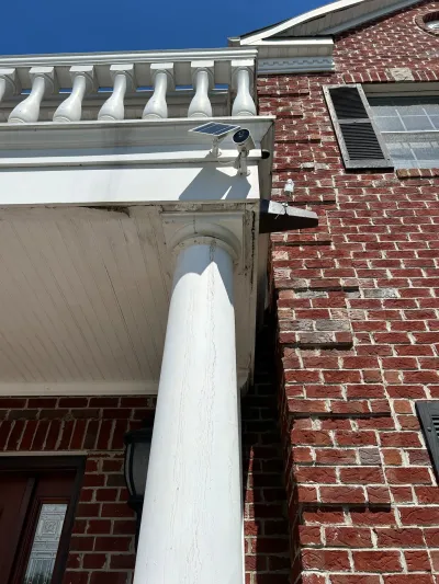 Close-up of a brick home's white column and porch trim with a solar security camera mounted overhead.