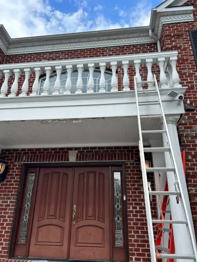 A ladder leans against a brick home's balcony above a double door entry with peeling porch ceiling paint.