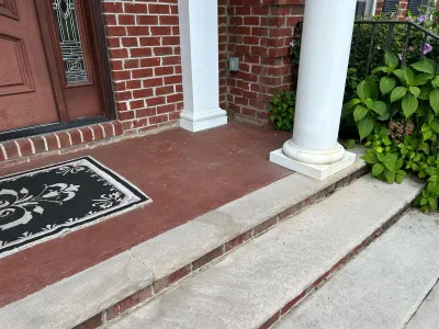 Cracked concrete porch floor and brick entry steps beside a white column and front door.