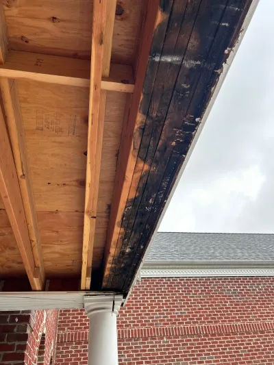 Close-up of new plywood framing next to a water-damaged, rotted wood porch overhang above a brick wall.
