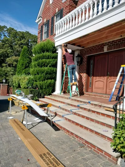A worker installs vinyl soffit on a ladder beneath the columned entry porch of a brick colonial home.