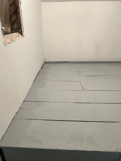 Empty room corner with grey-painted wood plank flooring and a wall access opening exposing insulation.