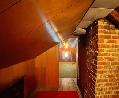 Narrow attic passageway with sloped wood paneling, an exposed brick chimney, and a lit room beyond.