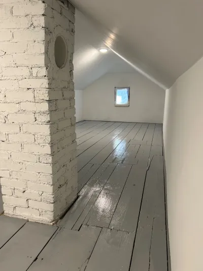 Empty attic room with a painted brick chimney, sloped ceiling, and grey painted wood plank flooring.