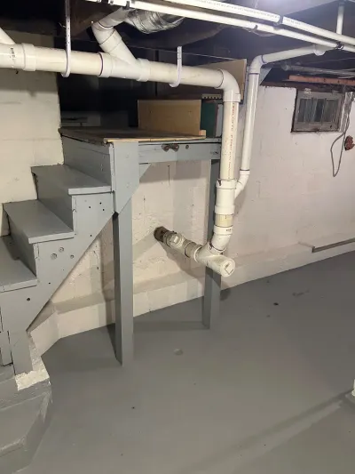 Unfinished basement with concrete block walls, PVC pipework, and a plywood storage platform under the stairs.